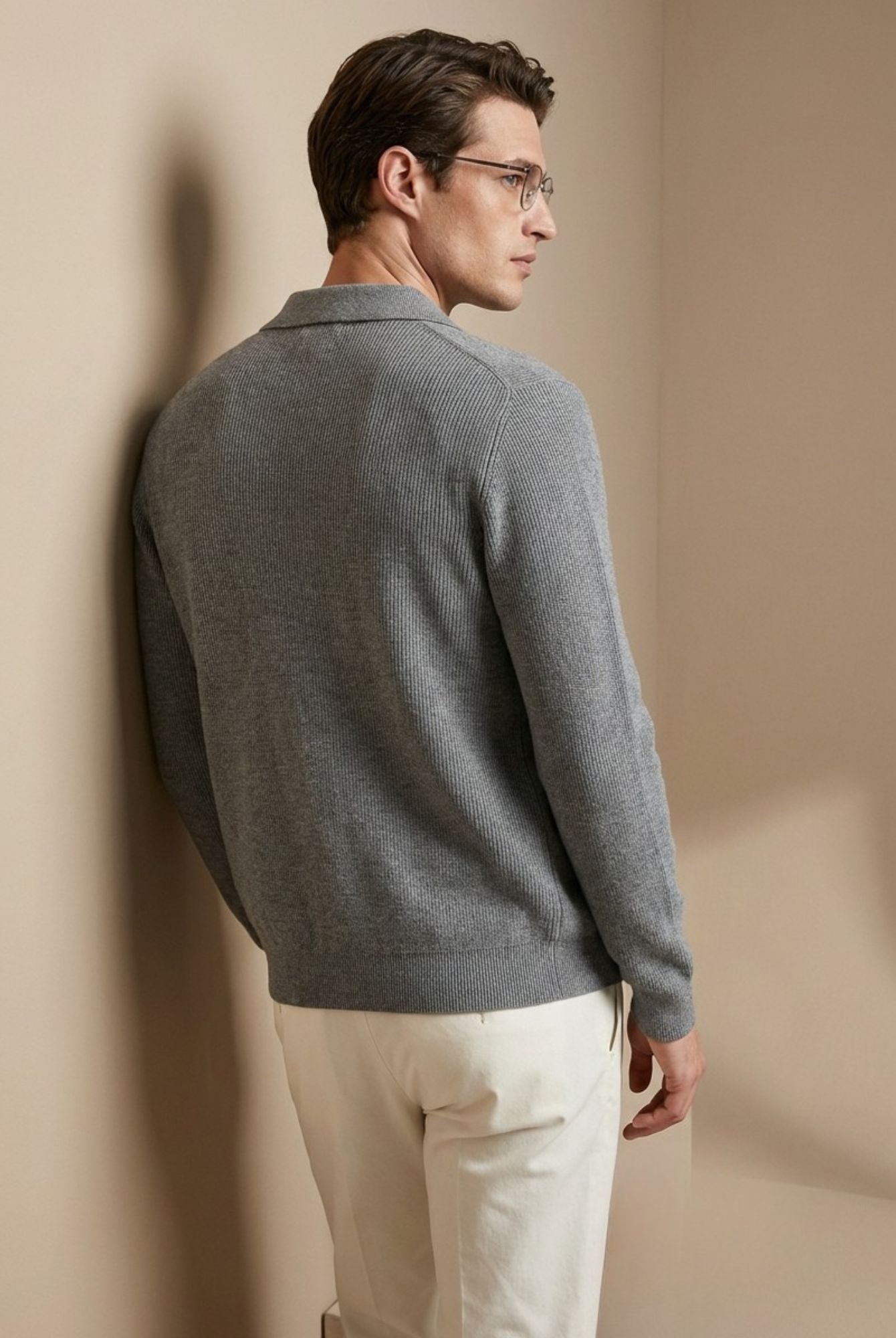 Man wearing a gray sweater and white pants against a beige wall