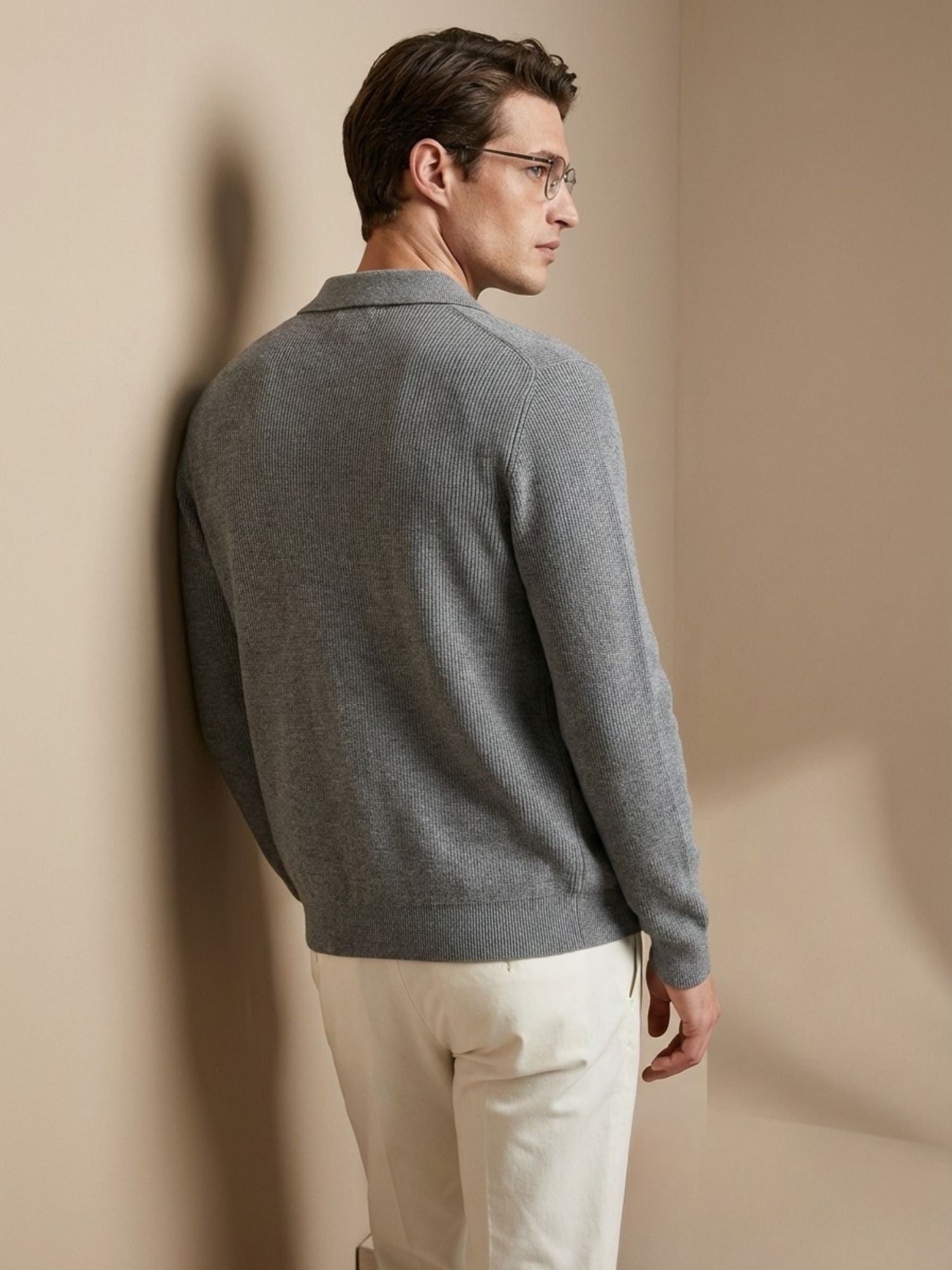 Man wearing a gray sweater and white pants against a beige wall