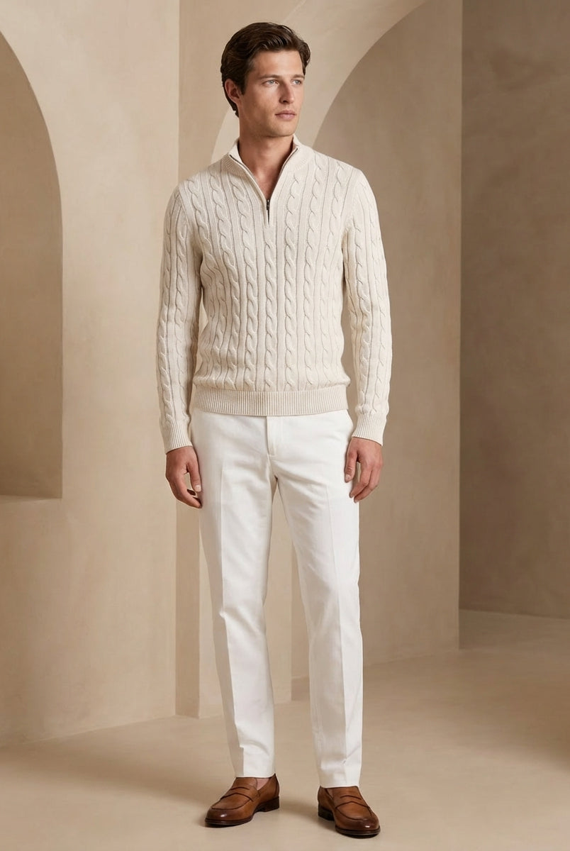 Man wearing a cream sweater and white pants standing in a neutral-colored room.