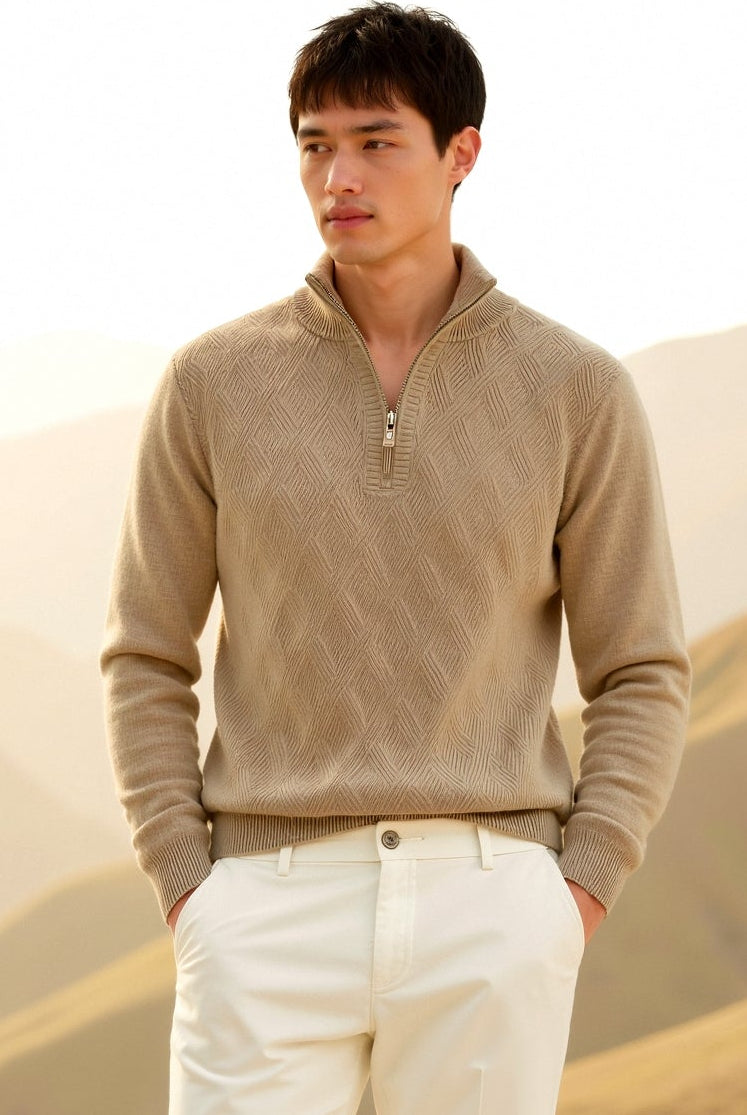 Man wearing a beige sweater and white pants standing in a desert landscape with mountains in the background.