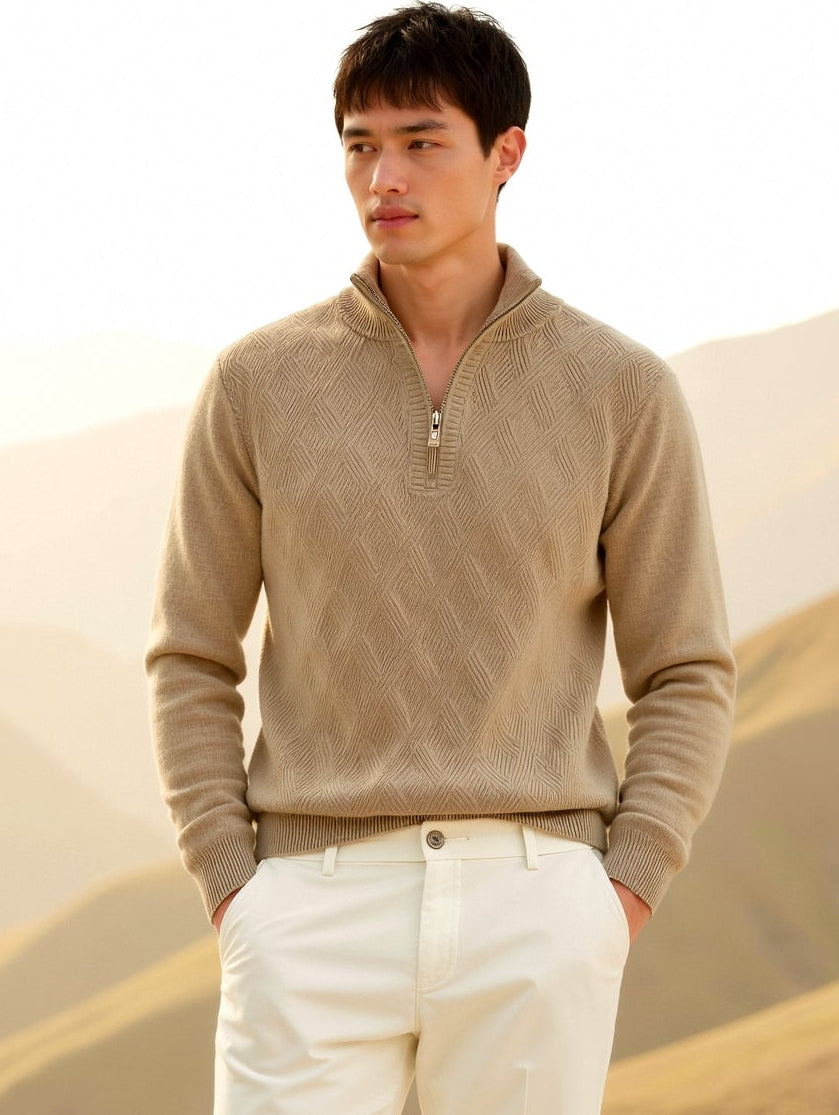 Man wearing a beige sweater and white pants standing in a desert landscape with mountains in the background.