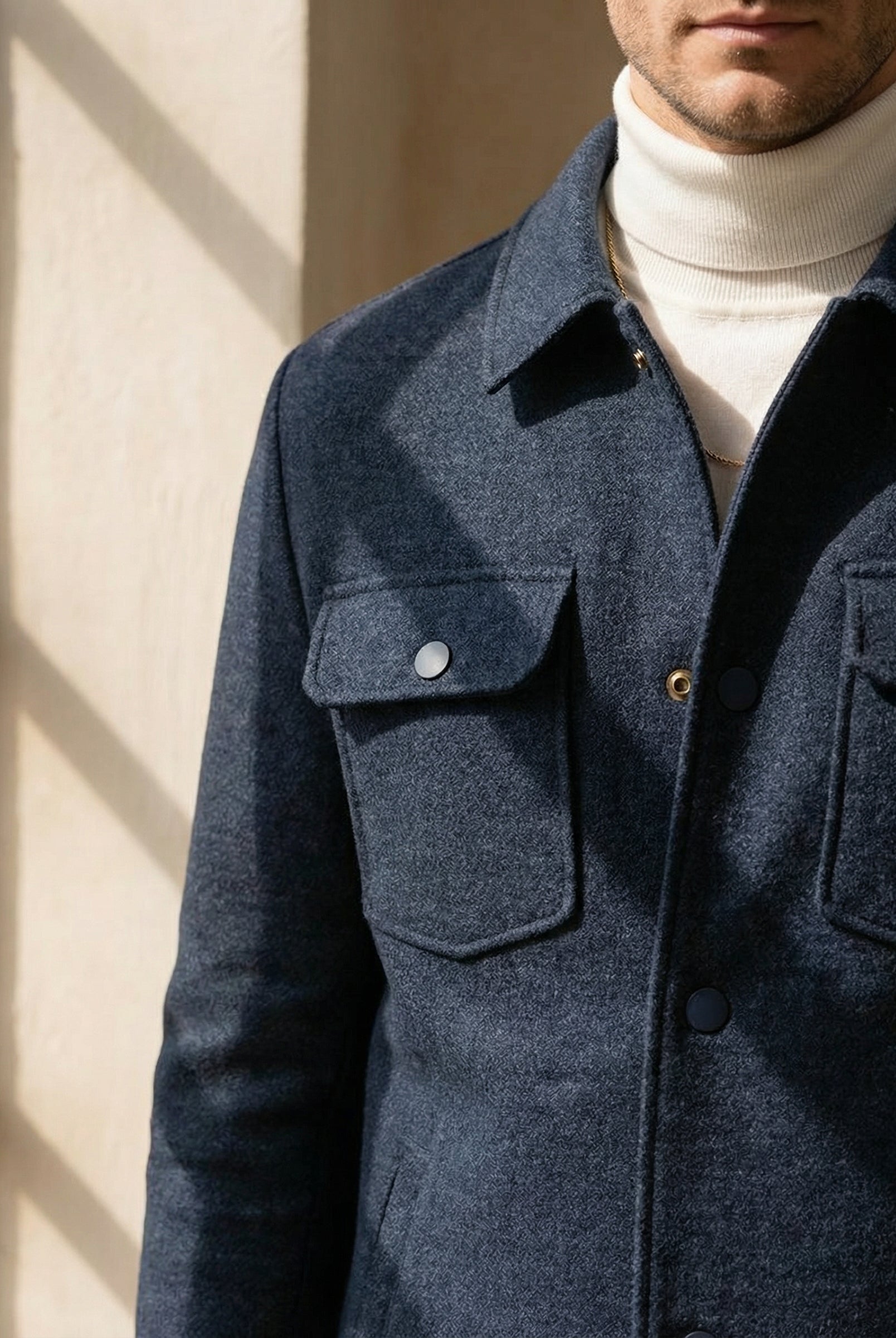 Man wearing a blue coat with a white turtleneck sweater, standing against a neutral background.