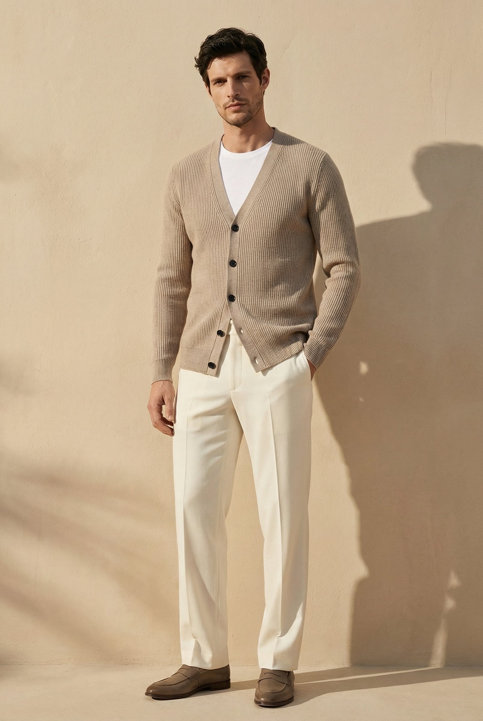 Man wearing a beige cardigan over a white shirt with white pants against a beige background