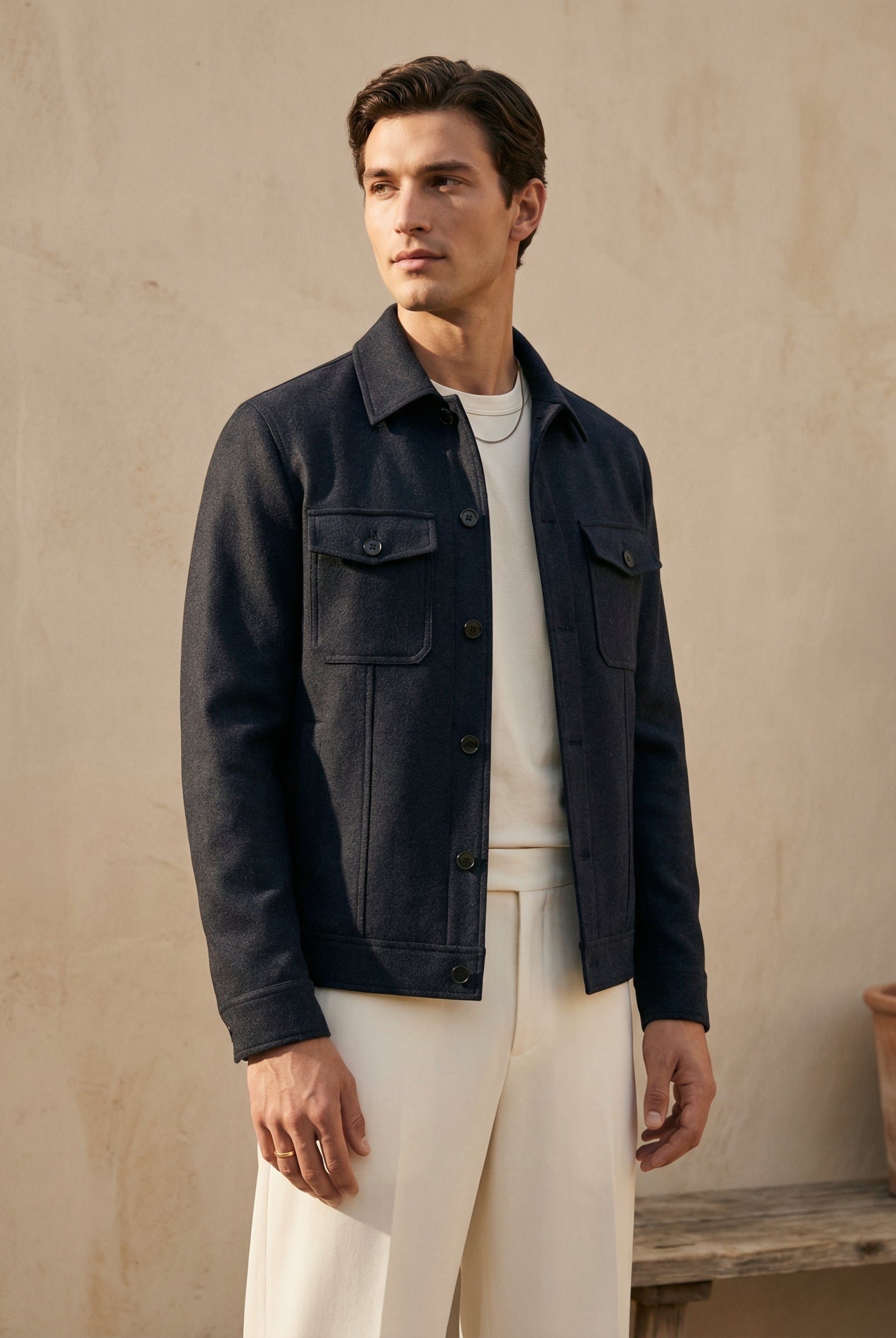Man wearing a dark jacket over a white shirt and pants against a beige wall.