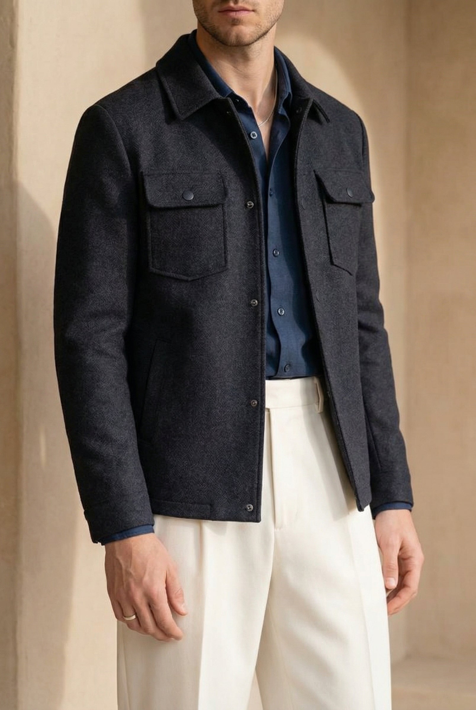 Man wearing a dark jacket over a blue shirt with white pants against a beige background