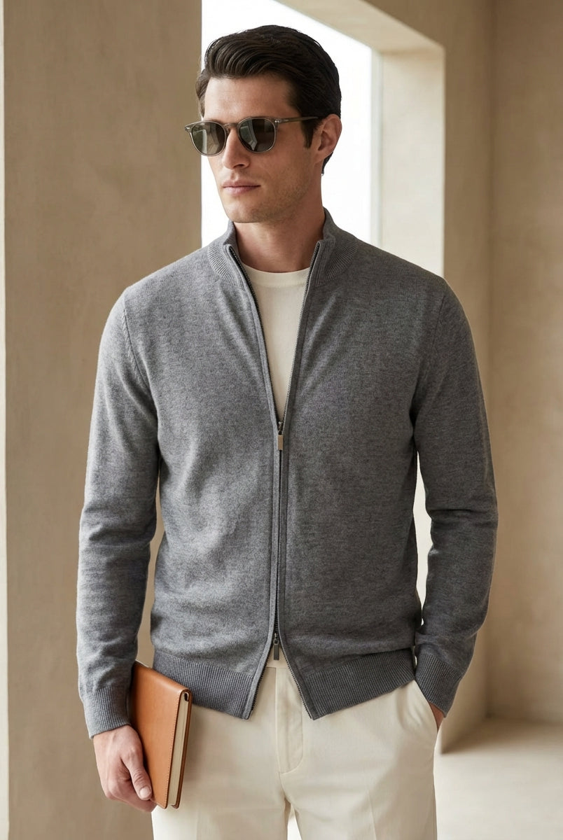 Man wearing a gray zip-up sweater and sunglasses, standing against a beige wall.