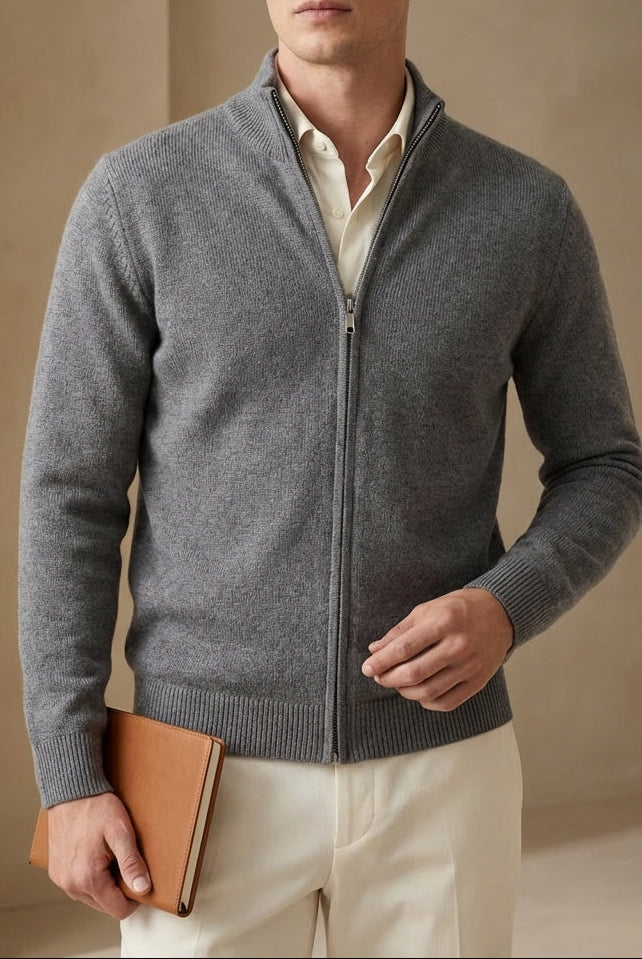 Man wearing a gray zip-up sweater over a white shirt, holding a brown leather jacket against a beige wall.
