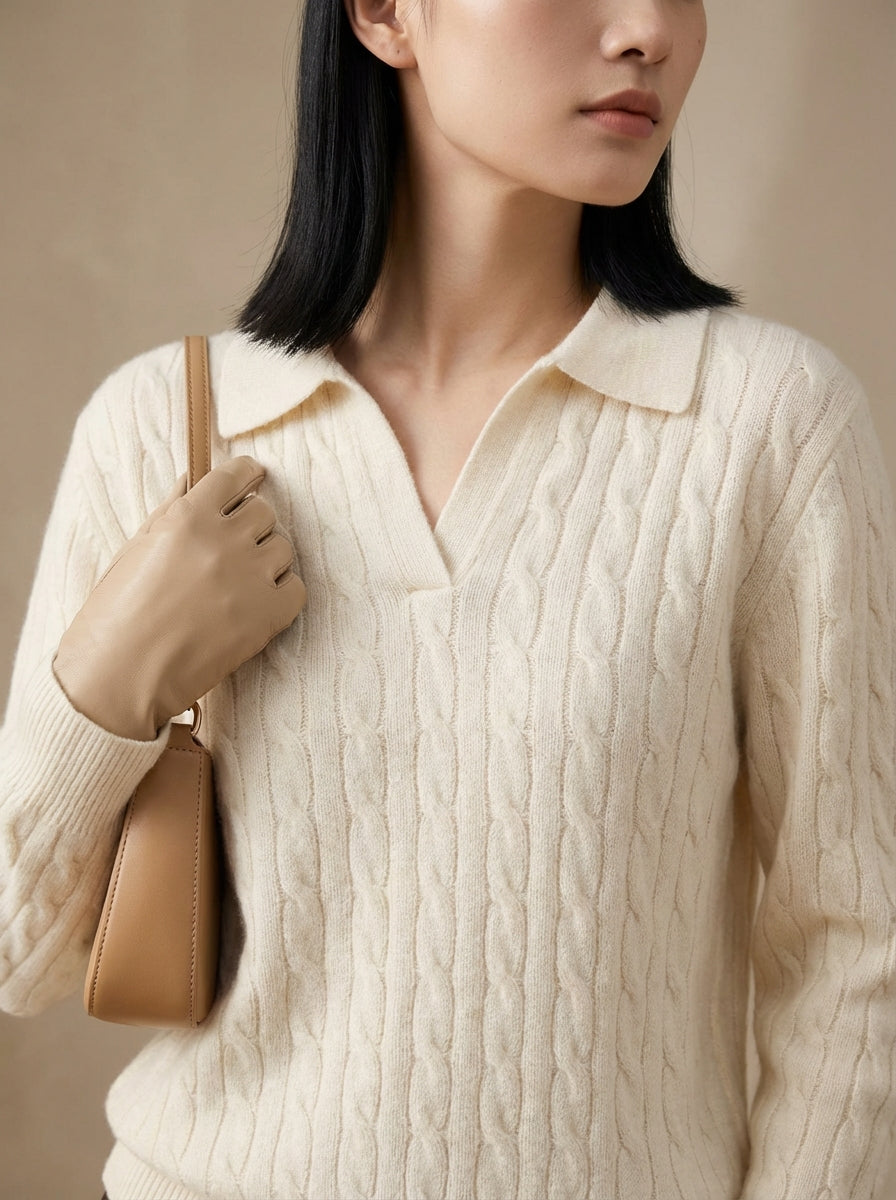 Woman wearing a cream cable knit sweater holding a beige handbag against a neutral background