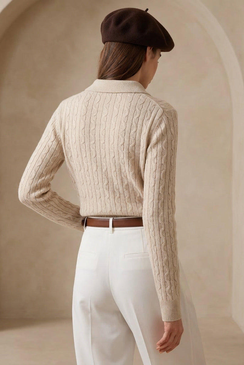 Person wearing a beige cable knit sweater and white pants with a brown beret, standing against a neutral background.