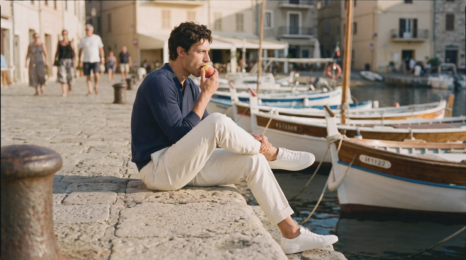 Man in a navy knitted polo, cream pleated trousers and plain white sneakers sitting on a harbour wall