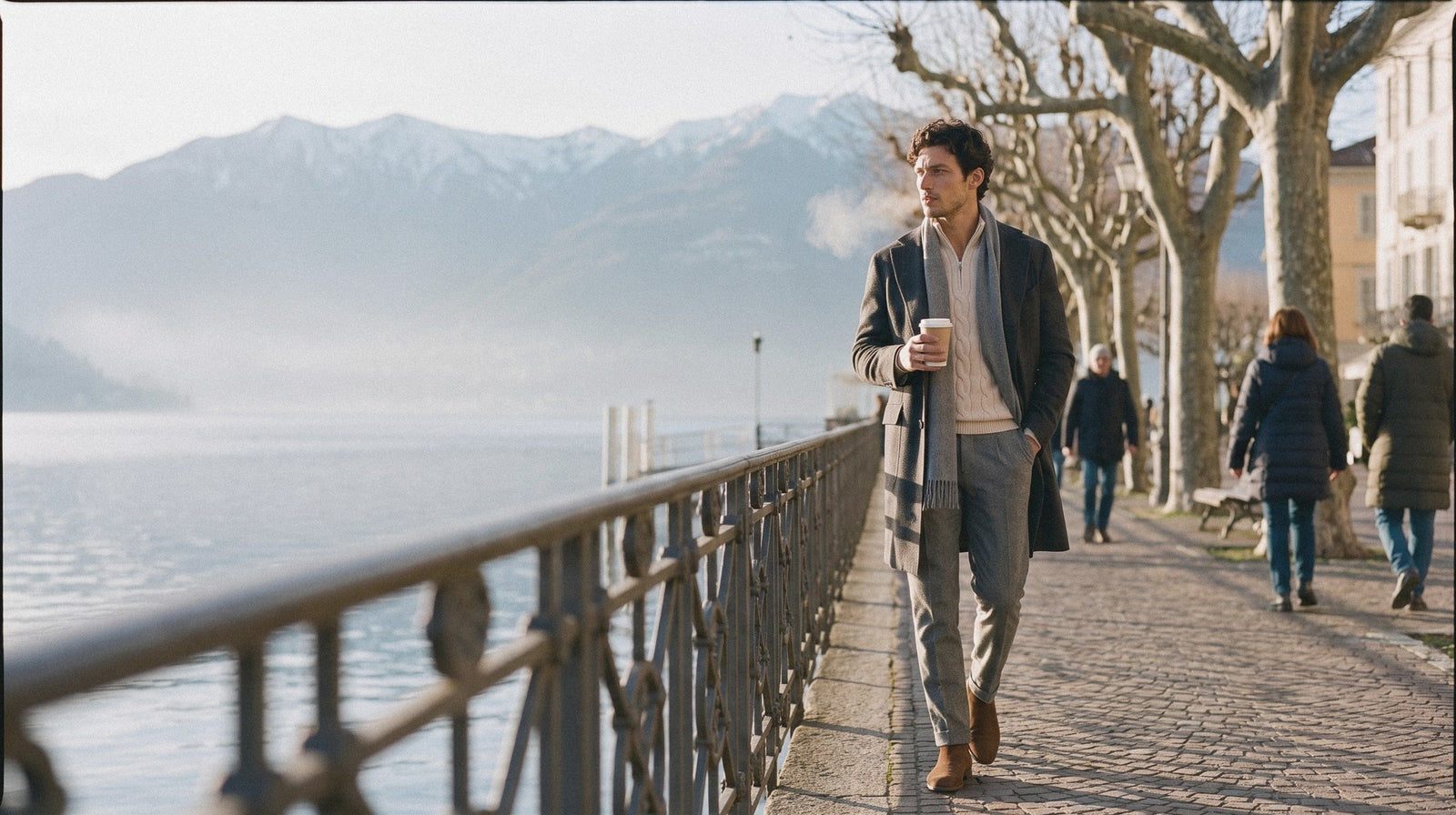 Man in a charcoal wool overcoat over a beige half zip cable knit walking by a lake on a cold winter morning