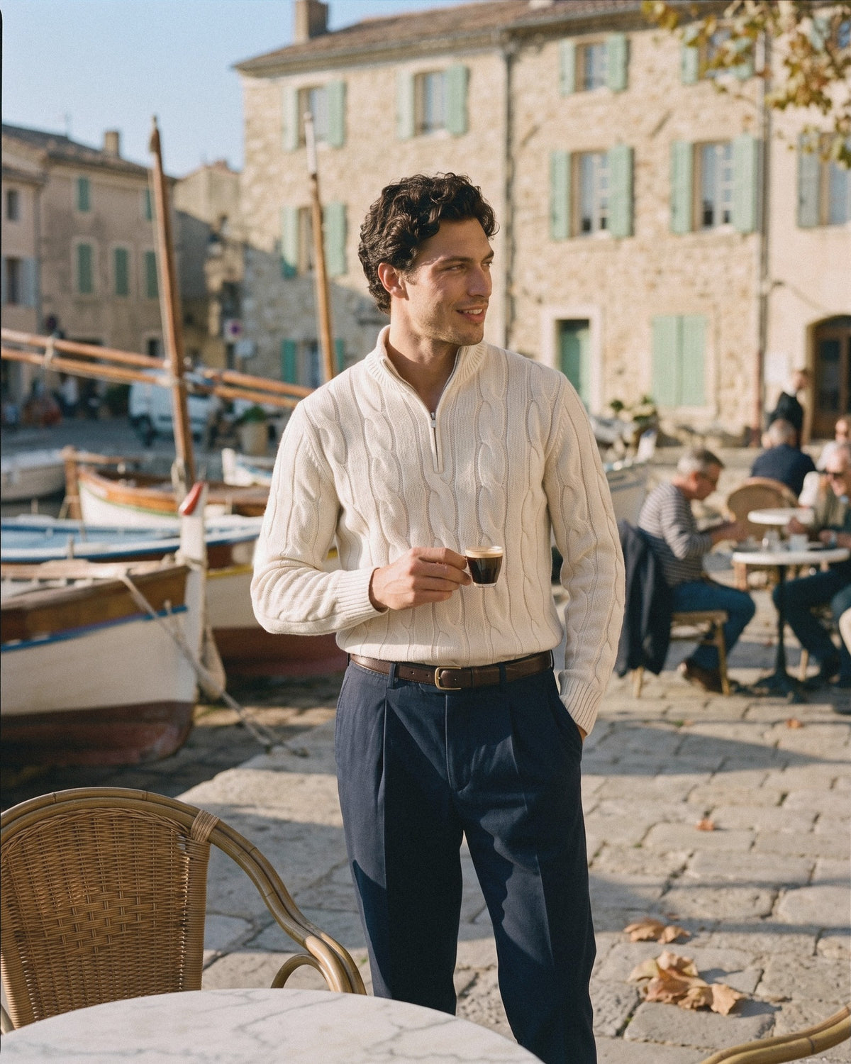 Man in an ivory cable-knit half-zip sweater and navy trousers at a harbour café in autumn