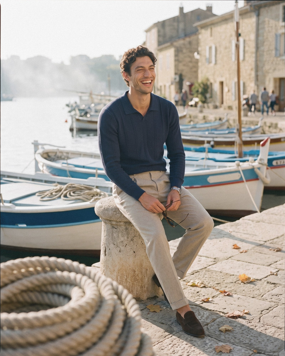 Man in a navy long-sleeve knit polo and beige trousers sitting on a harbour quay