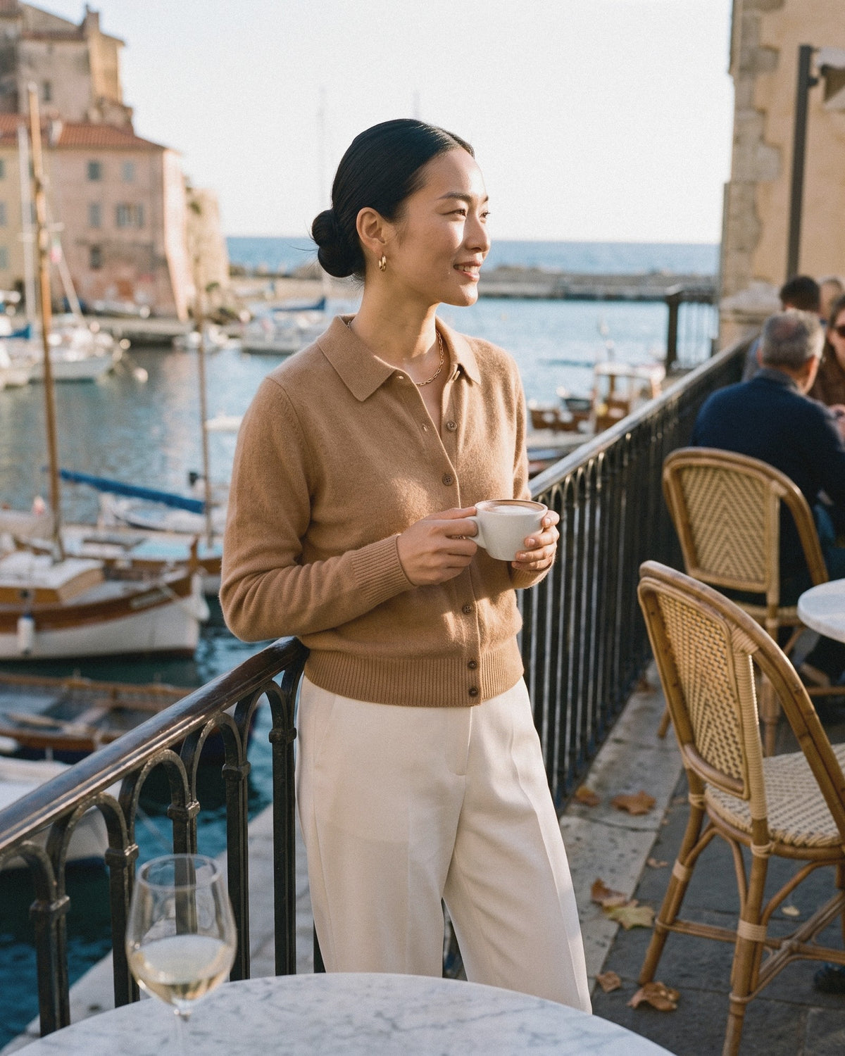 Woman in a camel knit polo cardigan and ivory trousers holding a coffee at a harbour café