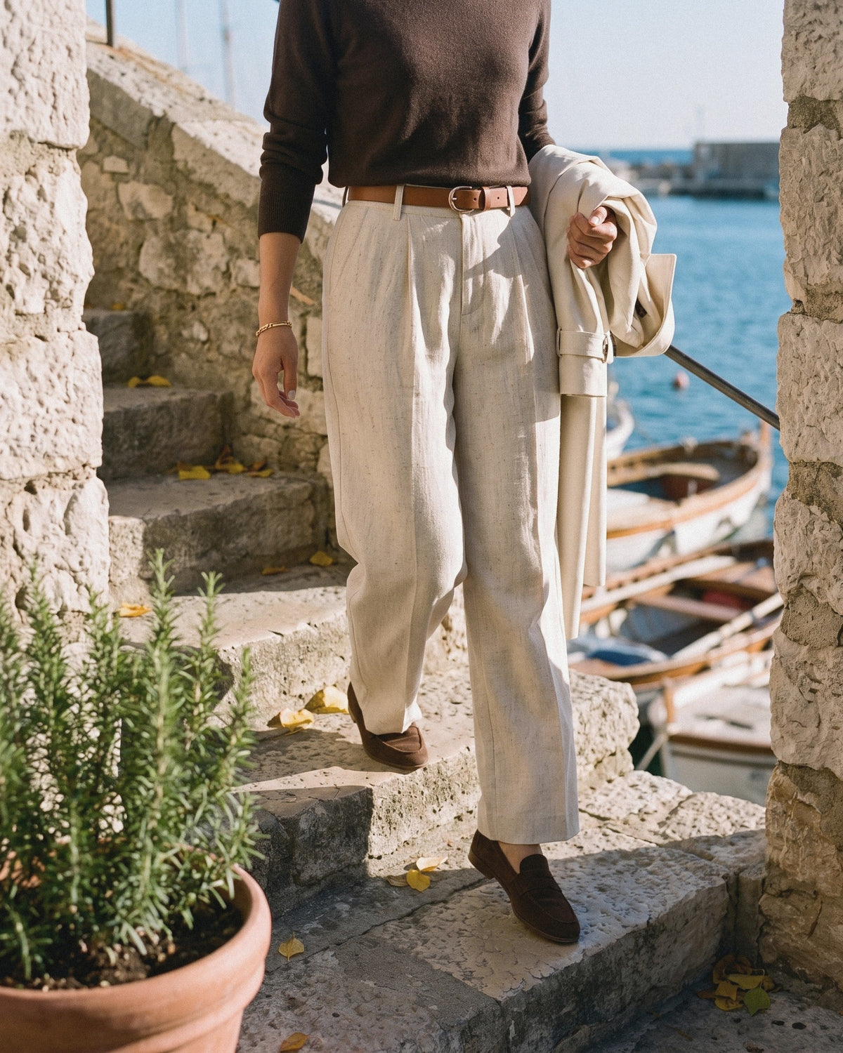 Woman in ivory wide-leg trousers and a brown knit walking down stone steps by the sea