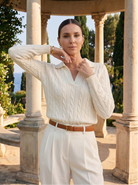 Woman in a white cable knit sweater and white pants standing in a scenic outdoor setting with columns and greenery.