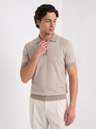 A man wearing a beige short-sleeve polo shirt with a subtle geometric pattern and cream trousers, standing against a plain white studio background.