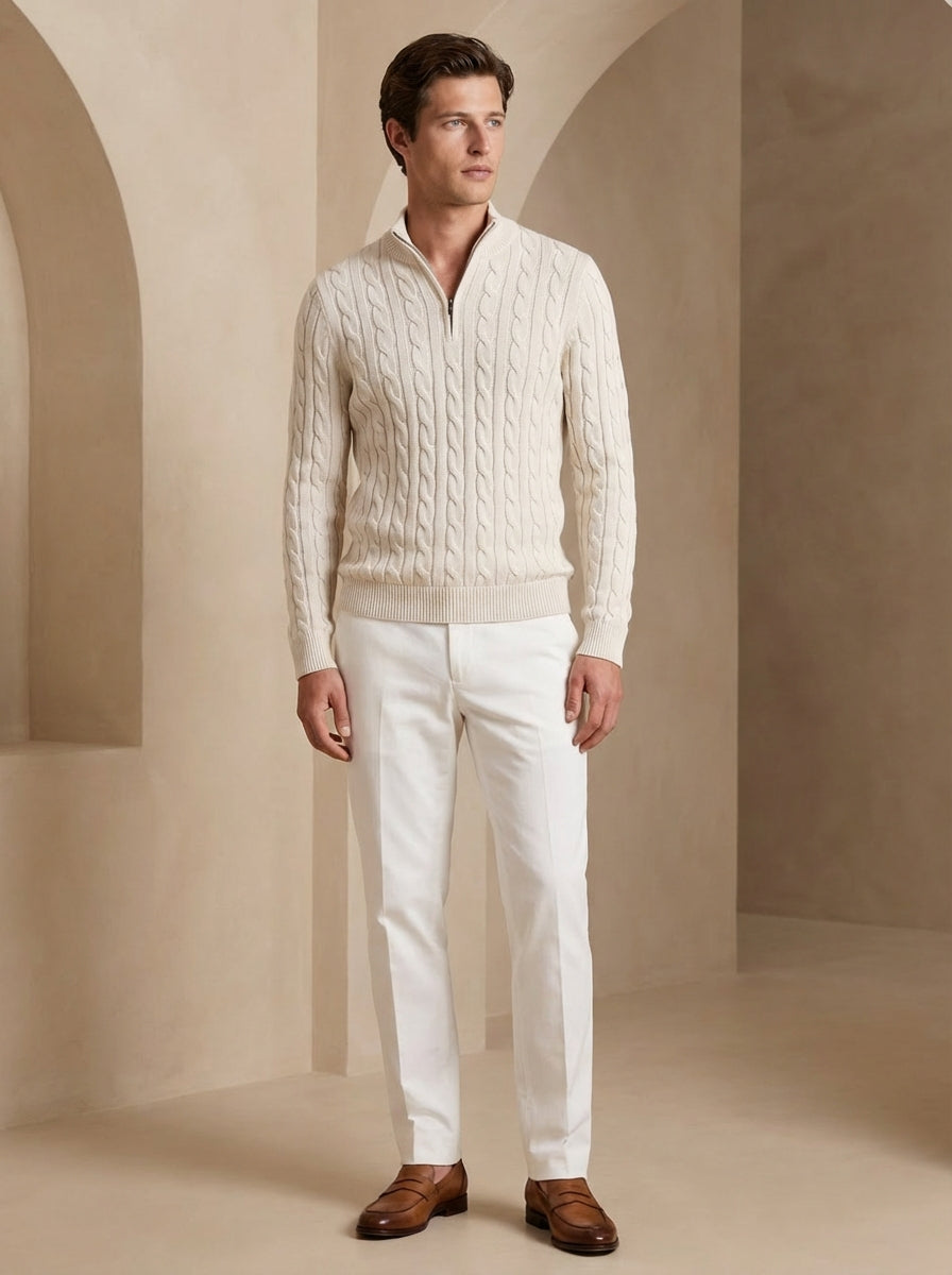 Man wearing a cream sweater and white pants standing in a neutral-colored room.