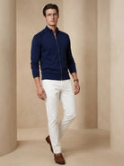 Man wearing a navy blue zip-up sweater, white pants, and brown shoes on a beige background