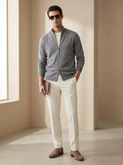 Man wearing a gray zip-up cardigan, white pants, and brown shoes in a minimalistic room.