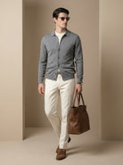 Man wearing a gray cardigan, white shirt, beige pants, and brown shoes, holding a brown leather bag against a beige background.