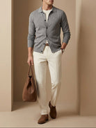 Man in gray cardigan and white pants holding a brown leather bag against a beige wall.
