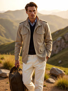 Man in beige jacket and white pants holding a suitcase against a mountainous landscape