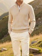 Man wearing a beige sweater and white pants standing in a mountainous landscape.