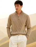Man wearing a beige sweater and white pants standing in a desert landscape with mountains in the background.