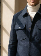 Man wearing a blue coat with a white turtleneck sweater, standing against a neutral background.