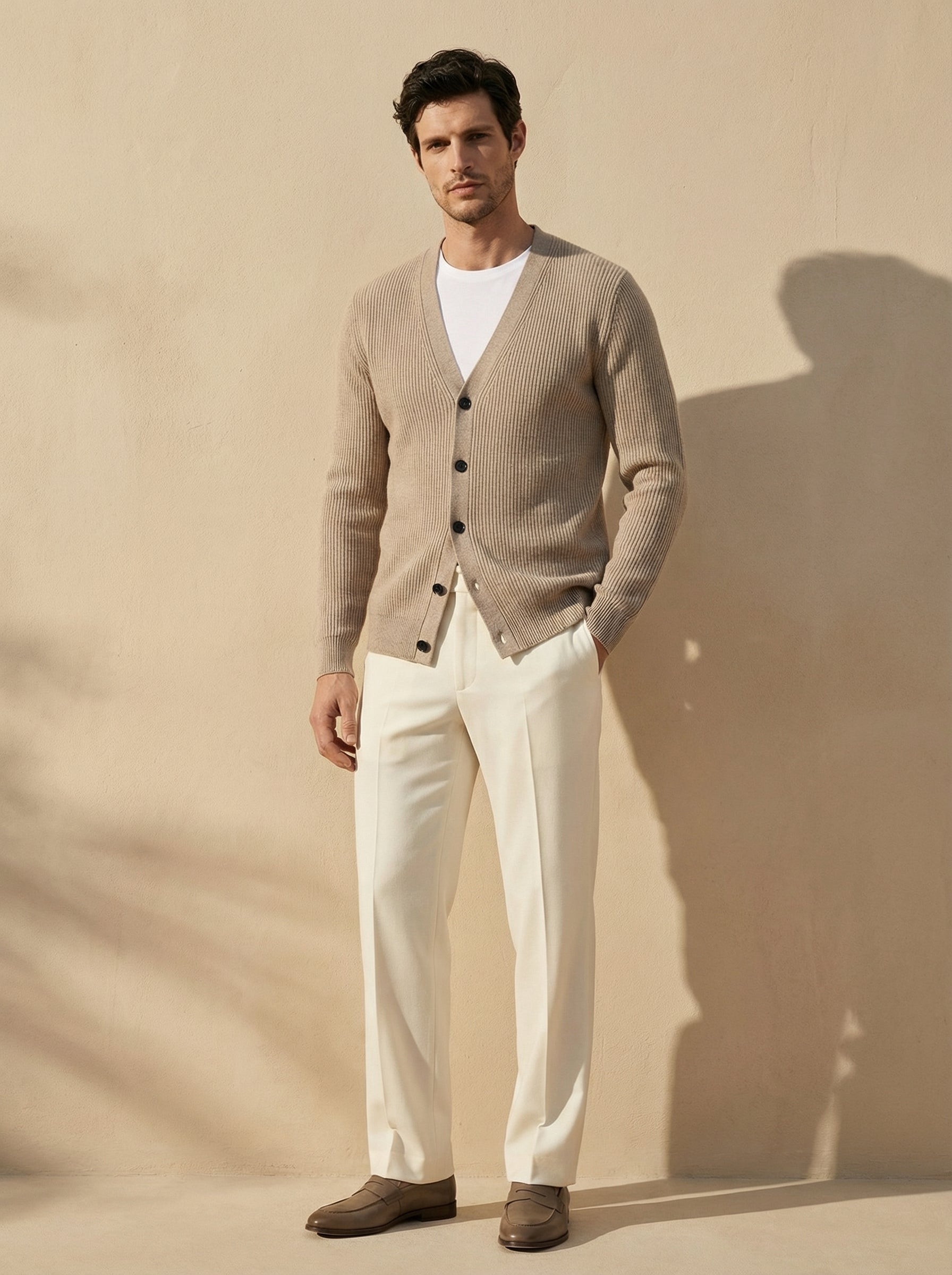Man wearing a beige cardigan over a white shirt with white pants against a beige background