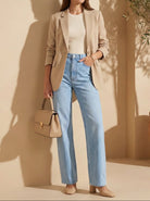 Woman in beige blazer and blue jeans holding a beige handbag against a beige wall.