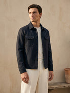 Man wearing a dark jacket over a white shirt and pants against a beige wall.