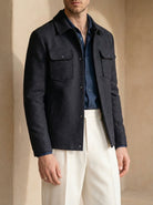 Man wearing a dark jacket over a blue shirt with white pants against a beige background