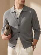 Man wearing a gray cardigan over a white shirt, holding a brown leather clutch against a beige background.