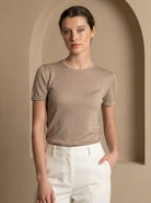 Woman wearing a brown t-shirt and white pants standing in a beige archway.