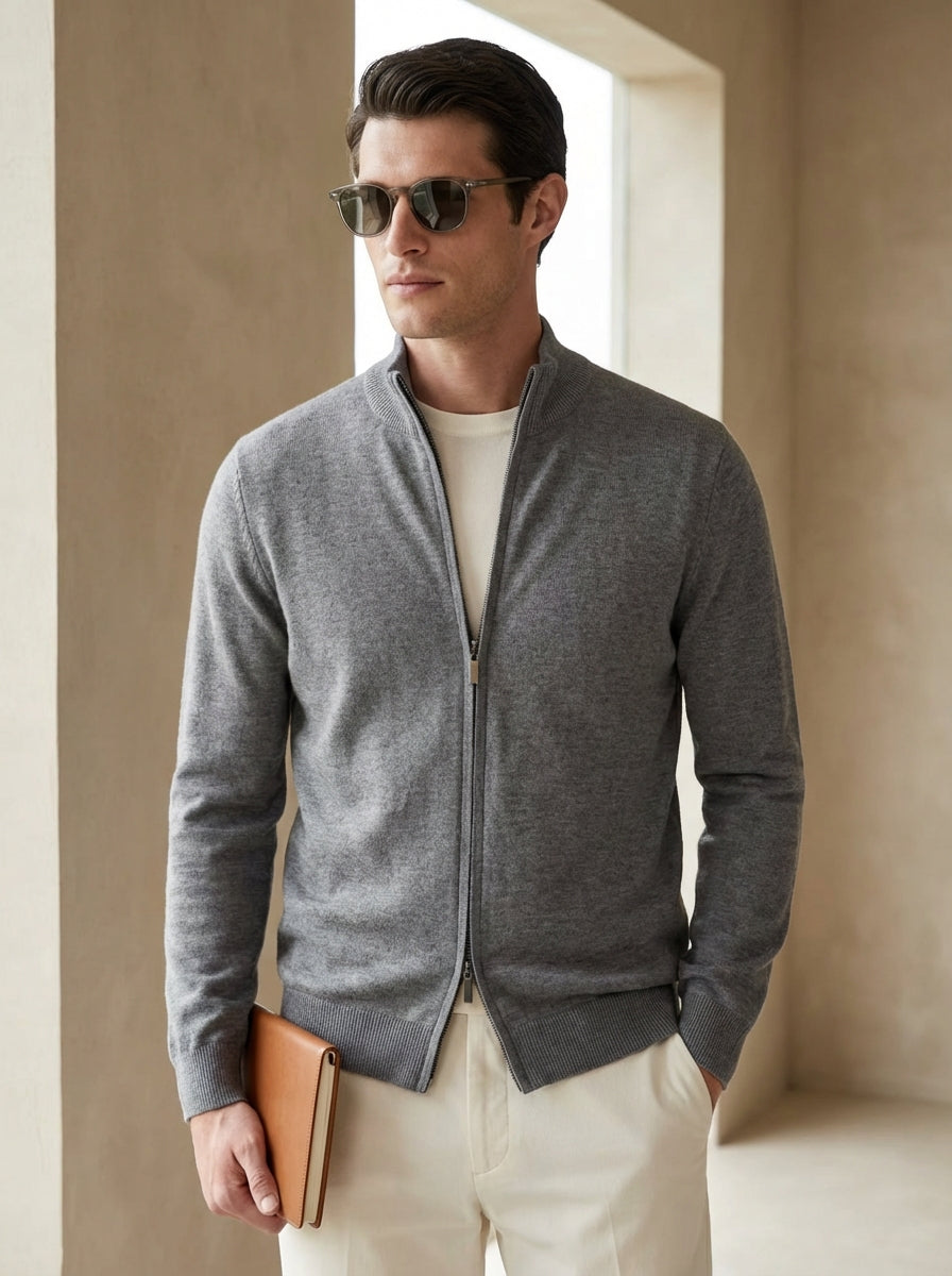 Man wearing a gray zip-up sweater and sunglasses, standing against a beige wall.