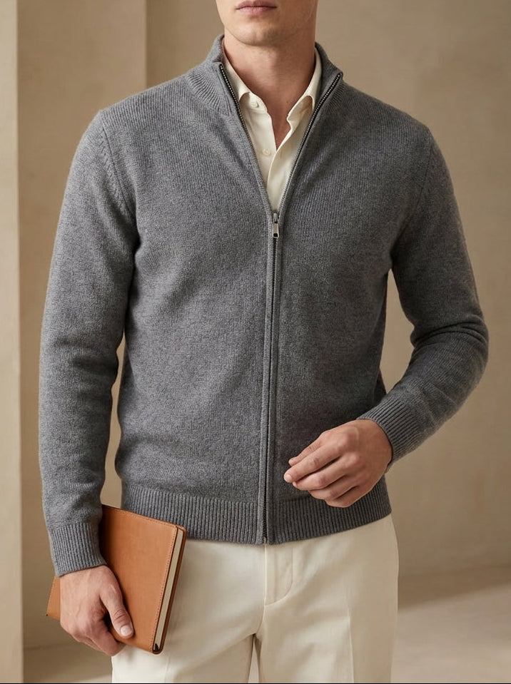 Man wearing a gray zip-up sweater over a white shirt, holding a brown leather jacket against a beige wall.