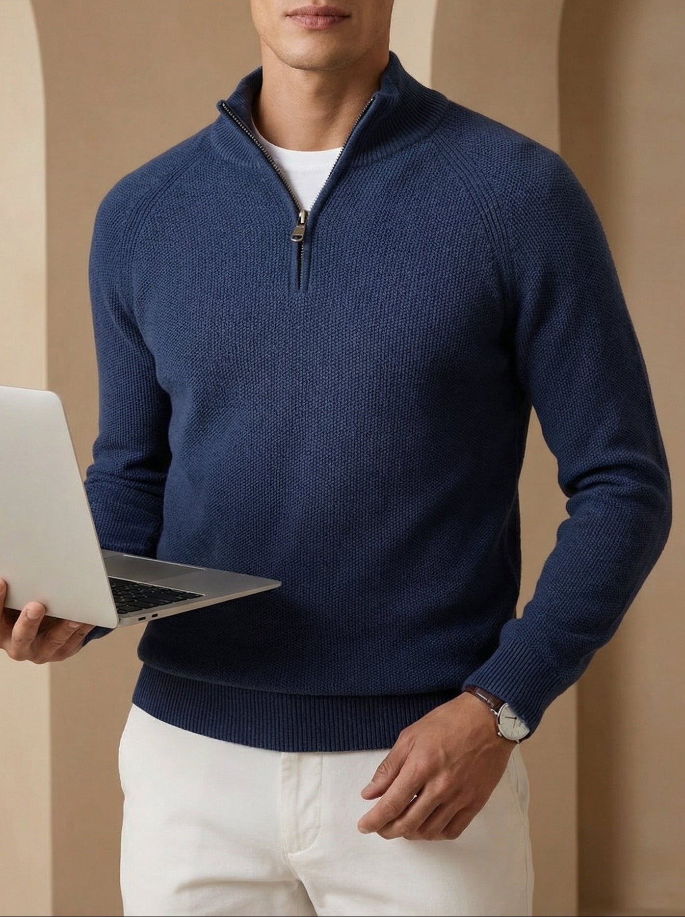 Man in a blue sweater holding a laptop against a beige archway background