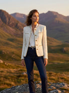 Woman standing in a mountainous landscape wearing a light-colored blazer and dark jeans.