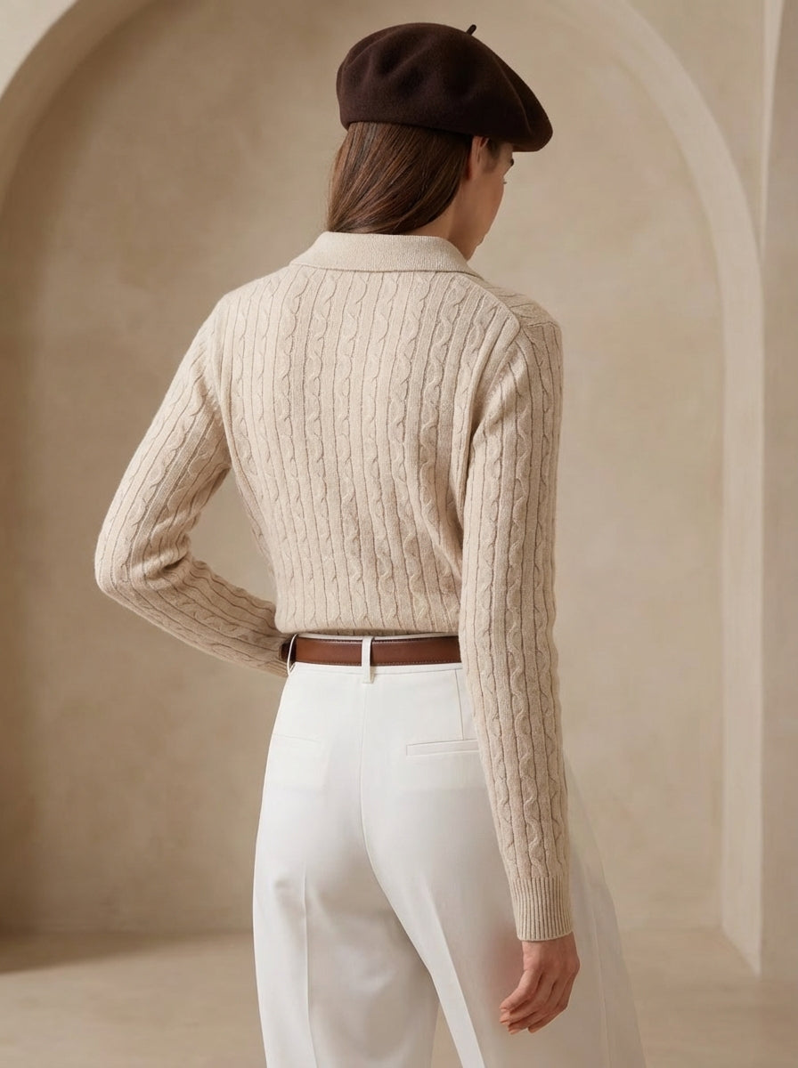 Person wearing a beige cable knit sweater and white pants with a brown beret, standing against a neutral background.