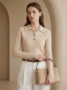 Woman wearing a beige cable knit sweater holding a matching handbag in an archway.