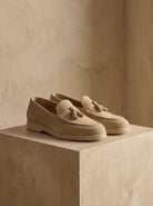 Pair of beige loafers with tassels on a beige platform against a neutral background